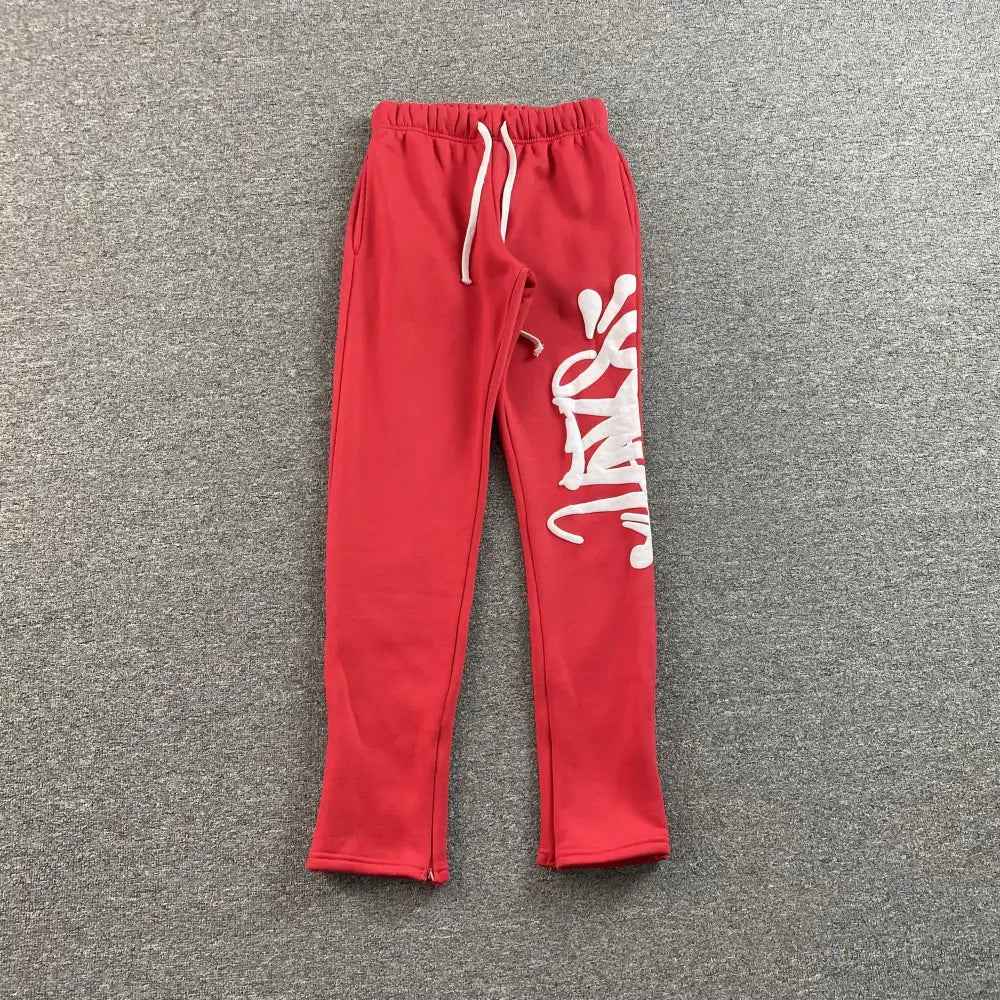 Red Syna World jogger pants with white graffiti letter print on right leg, laid flat