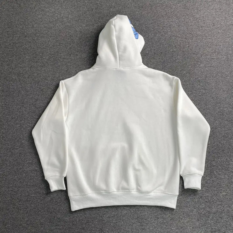 The Syna World White Blue Graffiti Letter Graphic Hoodie is laid flat, back facing up, on a gray textured surface. The hood is up and sleeves are spread out, showcasing the full streetwear design.