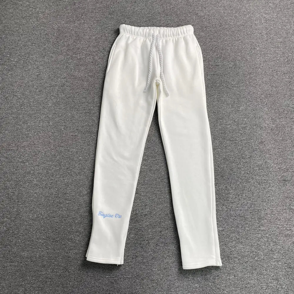 White Syna World jogger pants with light blue Forgive Me script embroidery on the lower left leg, laid flat on a grey surface