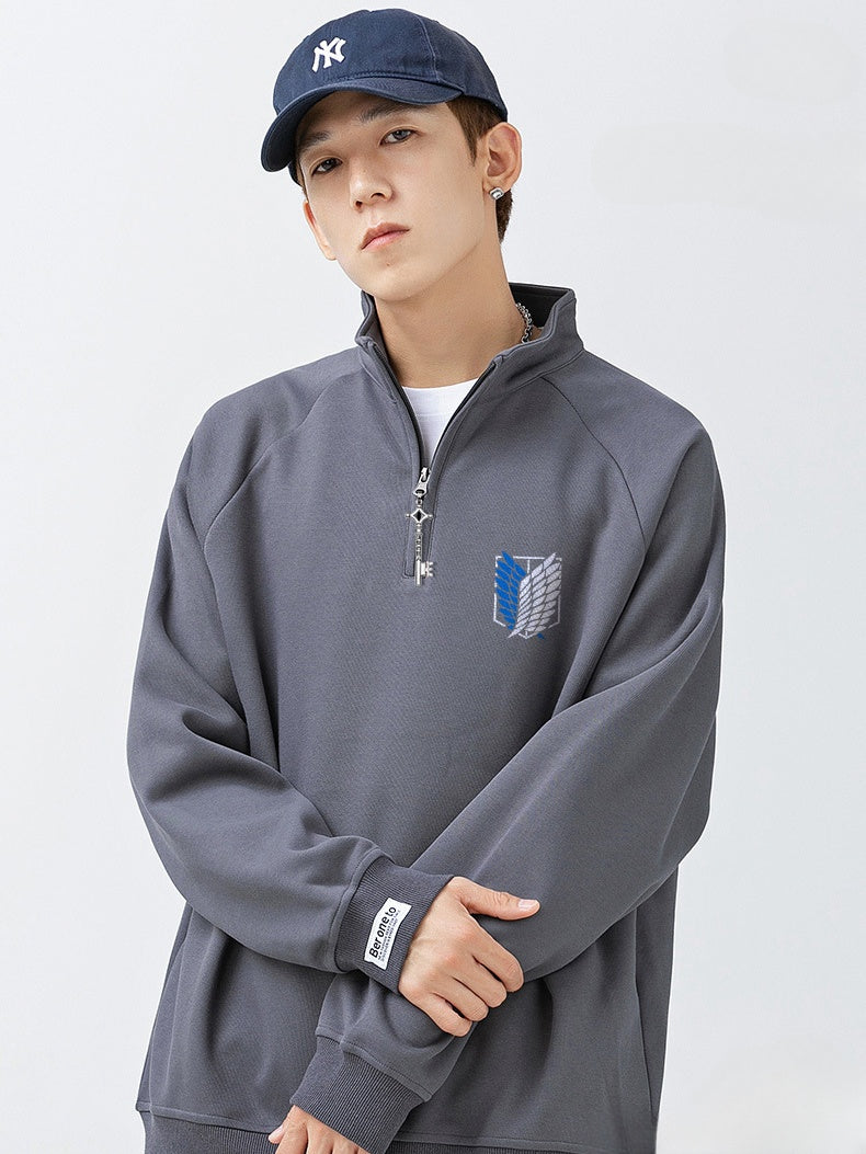 A person in a navy cap and gray Limited Edition Attack on Titan Half-Zip Sweatshirt stands against a light background. The sweatshirt showcases an embroidered Scout Regiment logo on the chest and Eren's Key zipper, subtly echoing the Wings of Freedom. The person gently crosses their arms.