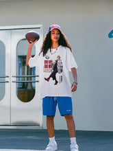 A person in a Jujutsu Kaisen Yuji Itadori NASA T-Shirt, blue shorts, and a baseball cap holds a football, standing outside near arched glass doors.